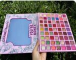 A large makeup kit displaying 63 square pans of highly pigmented eyeshadows, showcasing vibrant shades of blues, teals, greens, and purples in matte, shimmer, and glitter finishes.